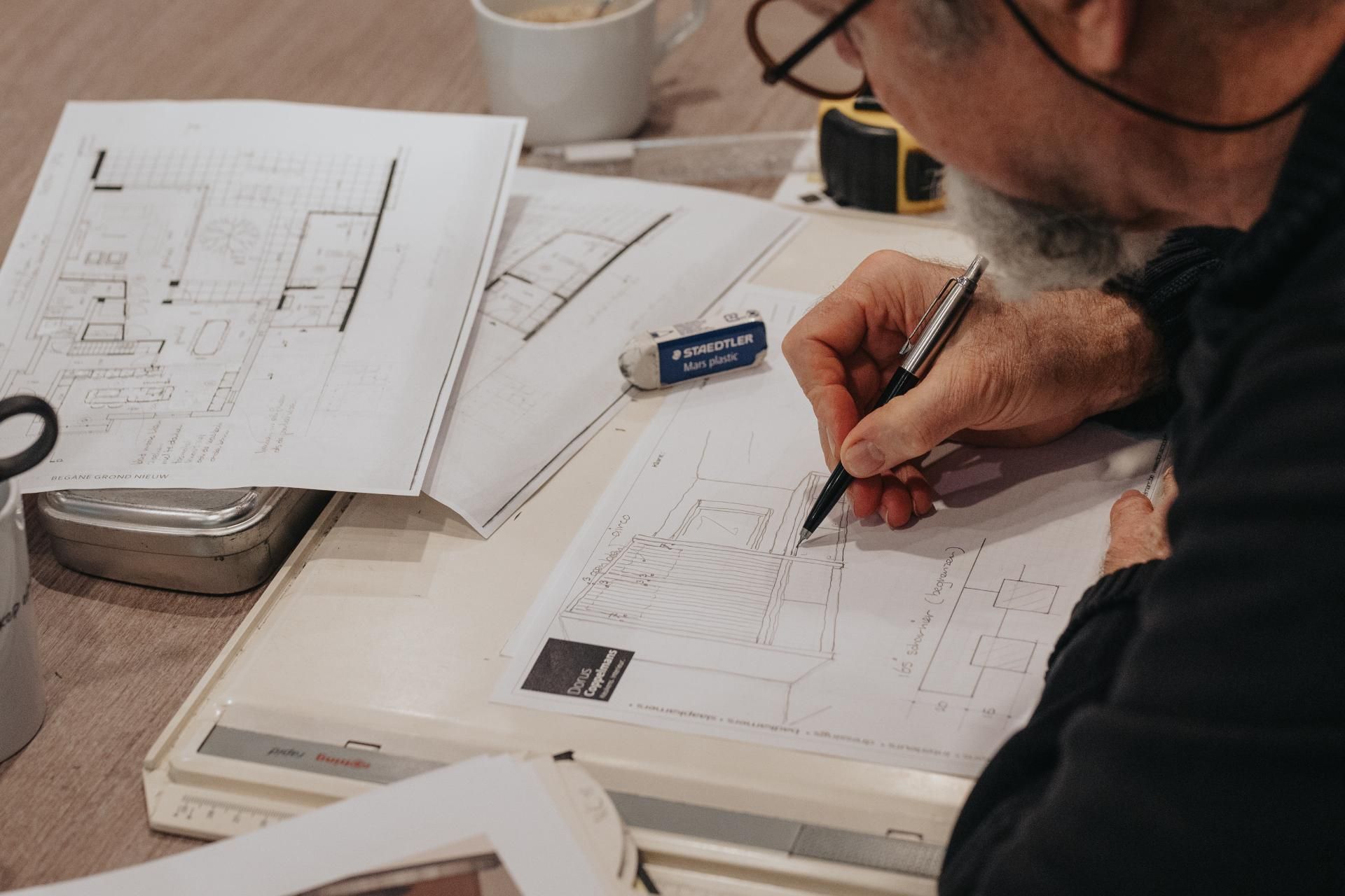 Een vakman tekent een interieurontwerp met de hand, omringd door technische bouwtekeningen en schetsen. Dit toont het ambachtelijke maatwerk en de precisie van Dorus Coppelmans in interieurdesign.