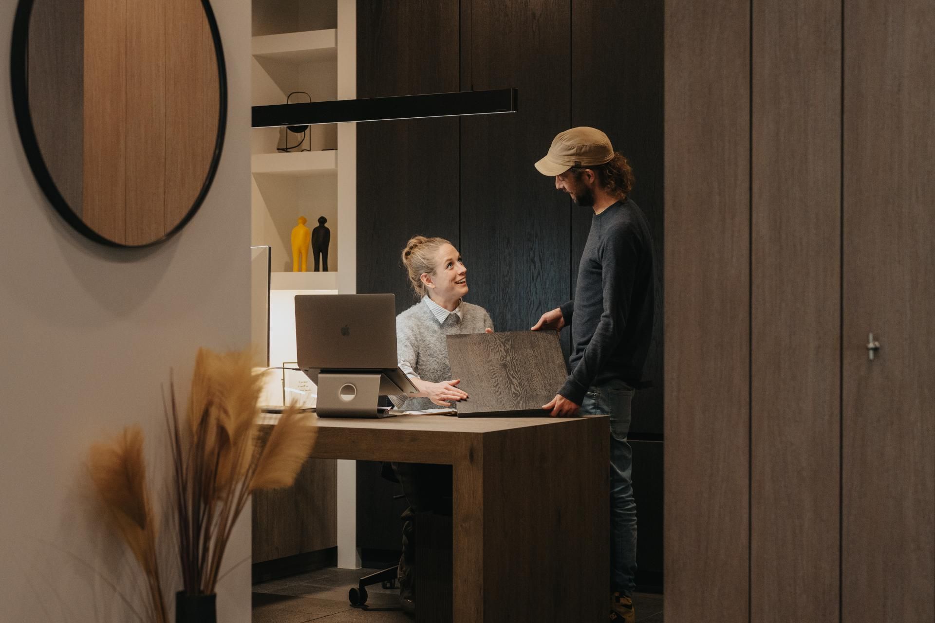 Een interieurontwerper zit aan een houten bureau en bespreekt een houtstaal met een klant in een stijlvolle showroom. De warme, moderne inrichting en de persoonlijke interactie benadrukken de focus op maatwerk interieuradvies.