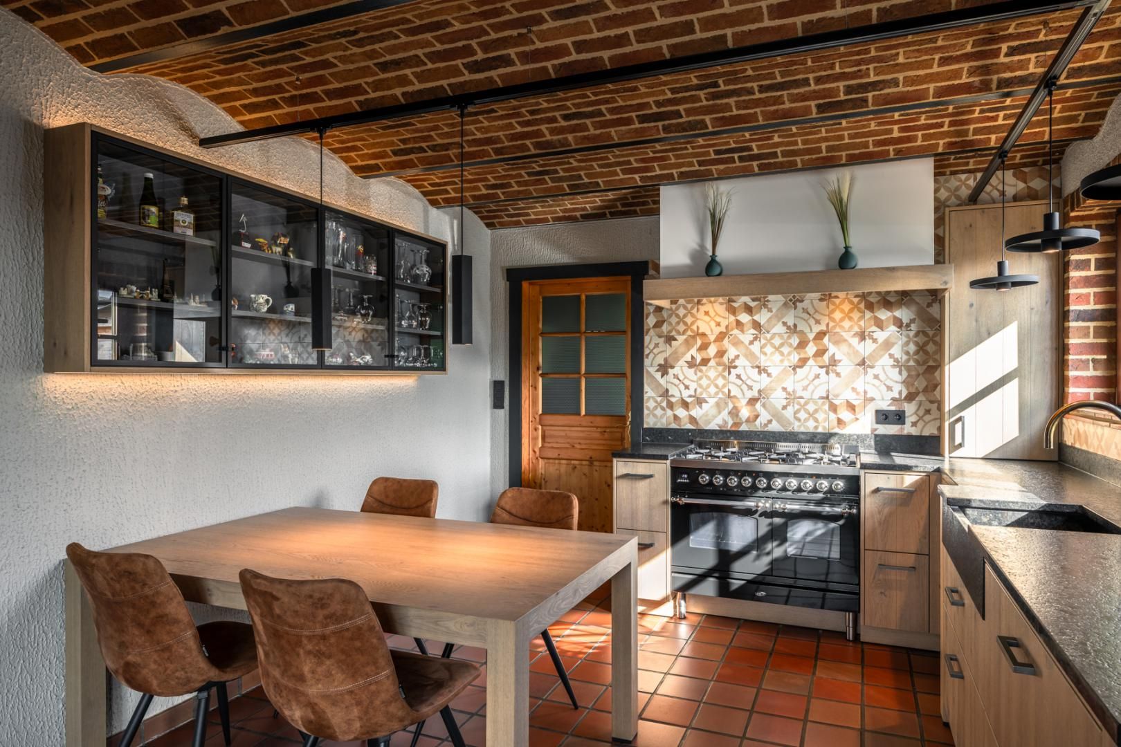 Gezellige landelijke keuken op maat met houten keukenkasten, een rustieke eettafel en lederen stoelen. De keuken combineert natuurlijke materialen met een sfeervolle bakstenen gewelvenplafond en een stijlvolle betegelde spatwand. Ambachtelijk vakmanschap en maatwerkinterieur zorgen voor een warme en functionele leefruimte.