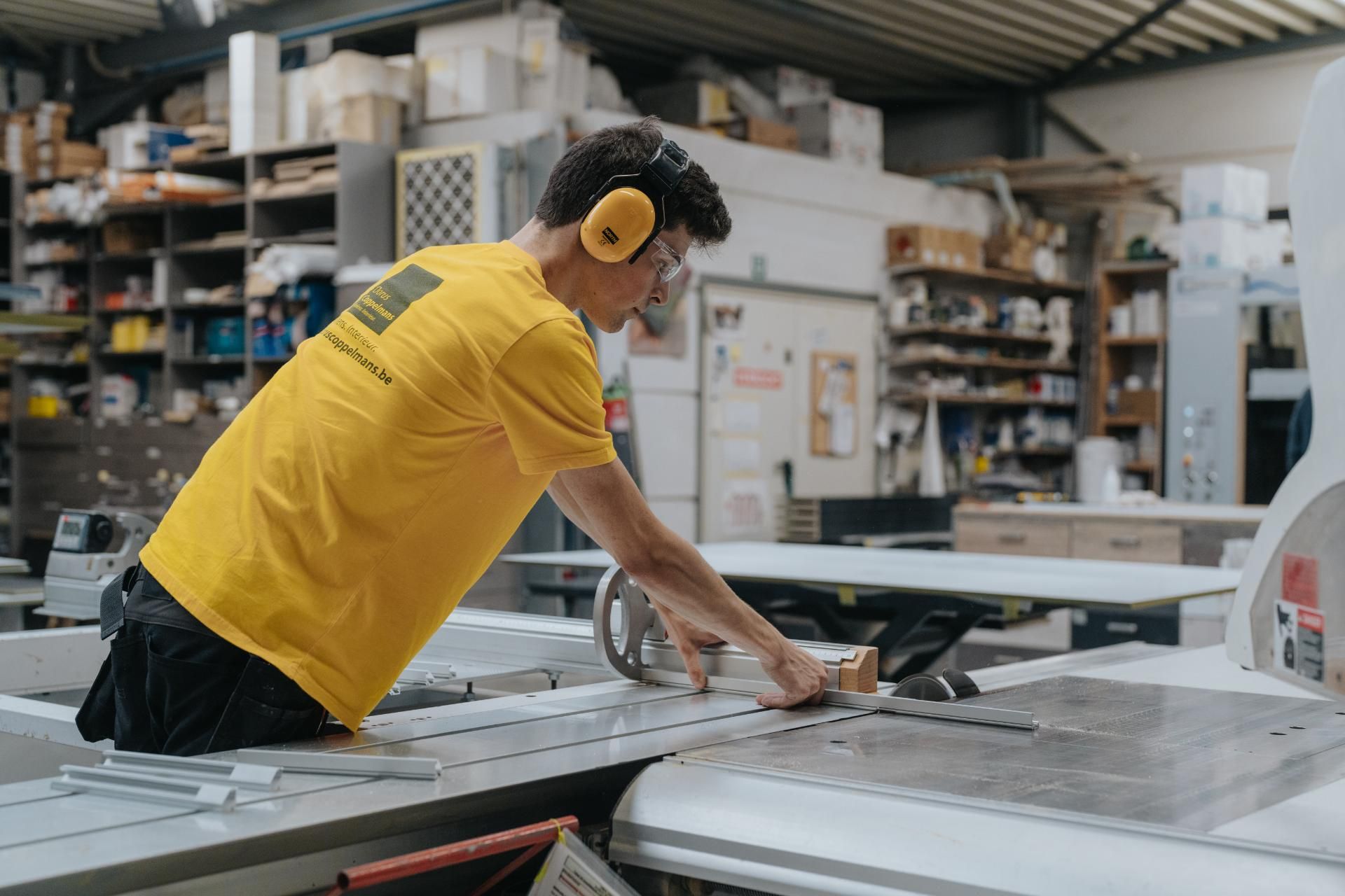 Een vakman in een gele werkshirt en gehoorbescherming werkt nauwkeurig aan een plaat hout op een zaagmachine in een ambachtelijke meubelmakerij. De werkplaats is gevuld met gereedschap en materialen, wat de precisie en kwaliteit van maatwerkinterieur benadrukt.