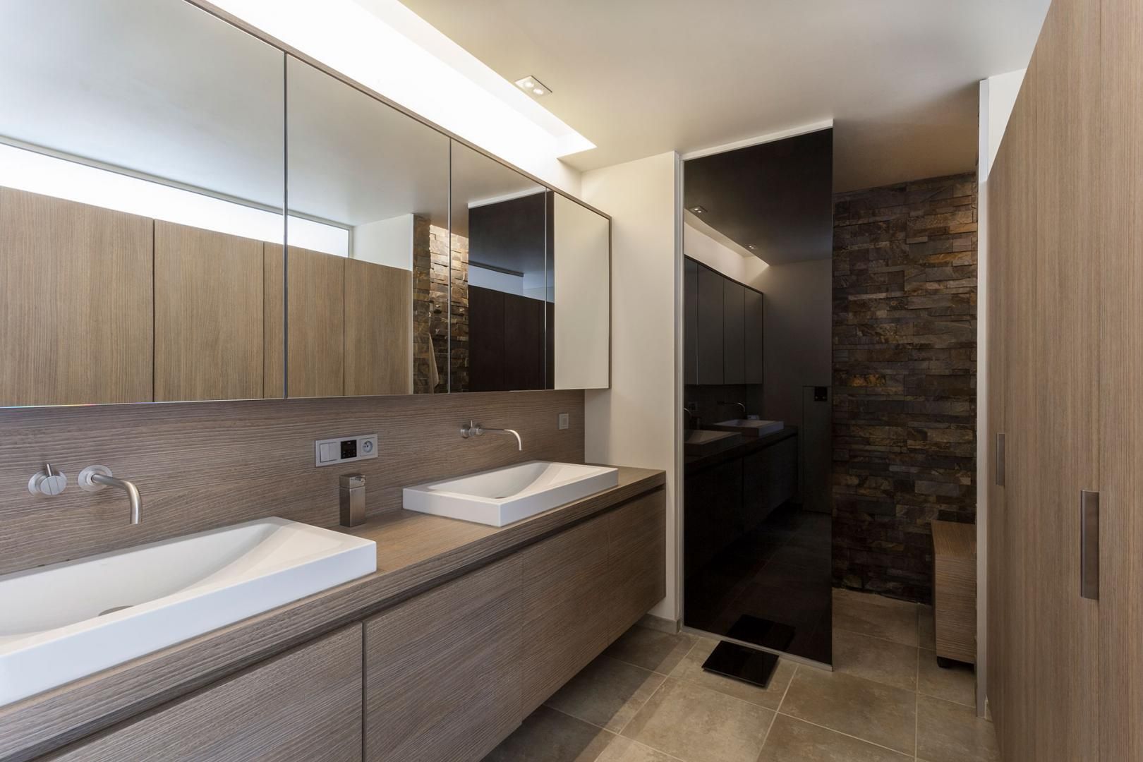 Een stijlvolle, moderne badkamer met een op maat gemaakt badkamermeubel, dubbele wastafels en grote spiegels. De combinatie van hout en natuursteen creëert een luxe spa-gevoel.