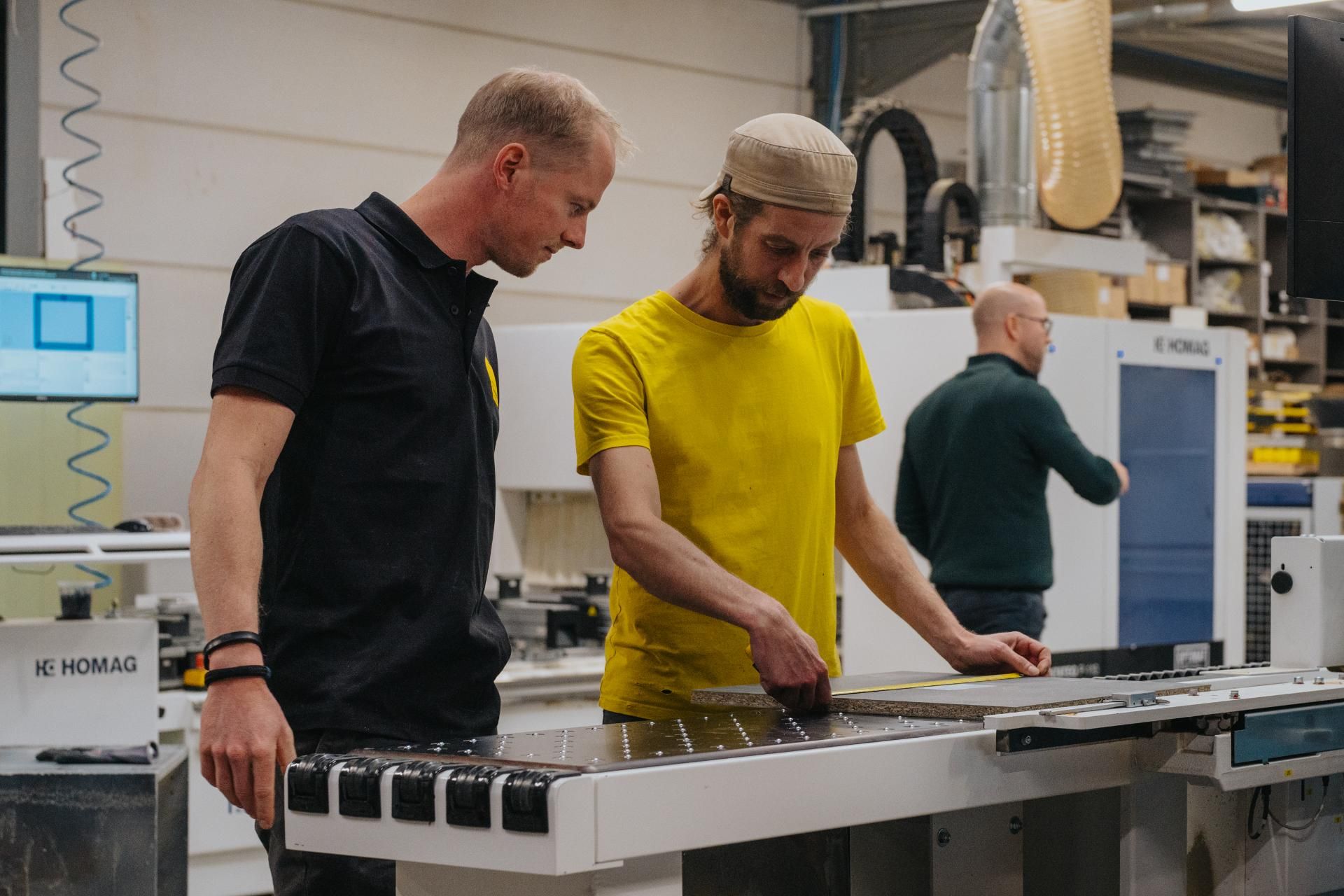 Twee vakmensen in een werkplaats meten en bewerken een houten plaat voor een op maat gemaakt interieurstuk. De combinatie van handwerk en geavanceerde machines benadrukt het ambachtelijke en duurzame vakmanschap.
