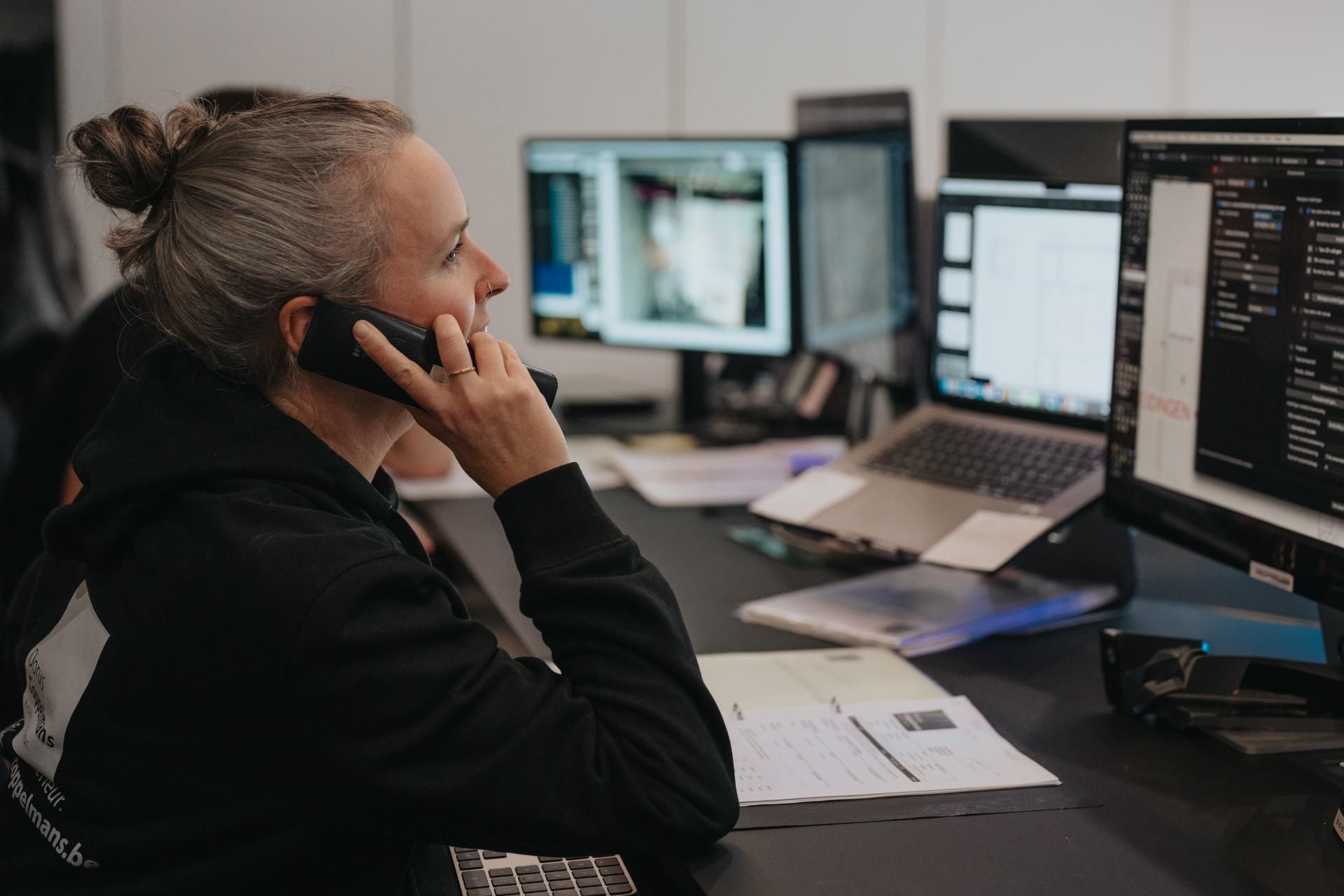 Een medewerker zit aan haar bureau en voert een telefoongesprek terwijl ze werkt aan interieurontwerpen op meerdere computerschermen. De professionele werkomgeving straalt expertise en nauwkeurigheid in maatwerkinterieurs uit.